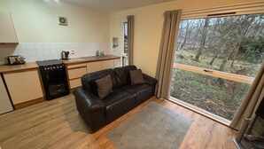 A compact living space with a kitchenette, a black leather sofa, and a large window offering a forest view. - Burnbank Lodges (Spean Bridge)