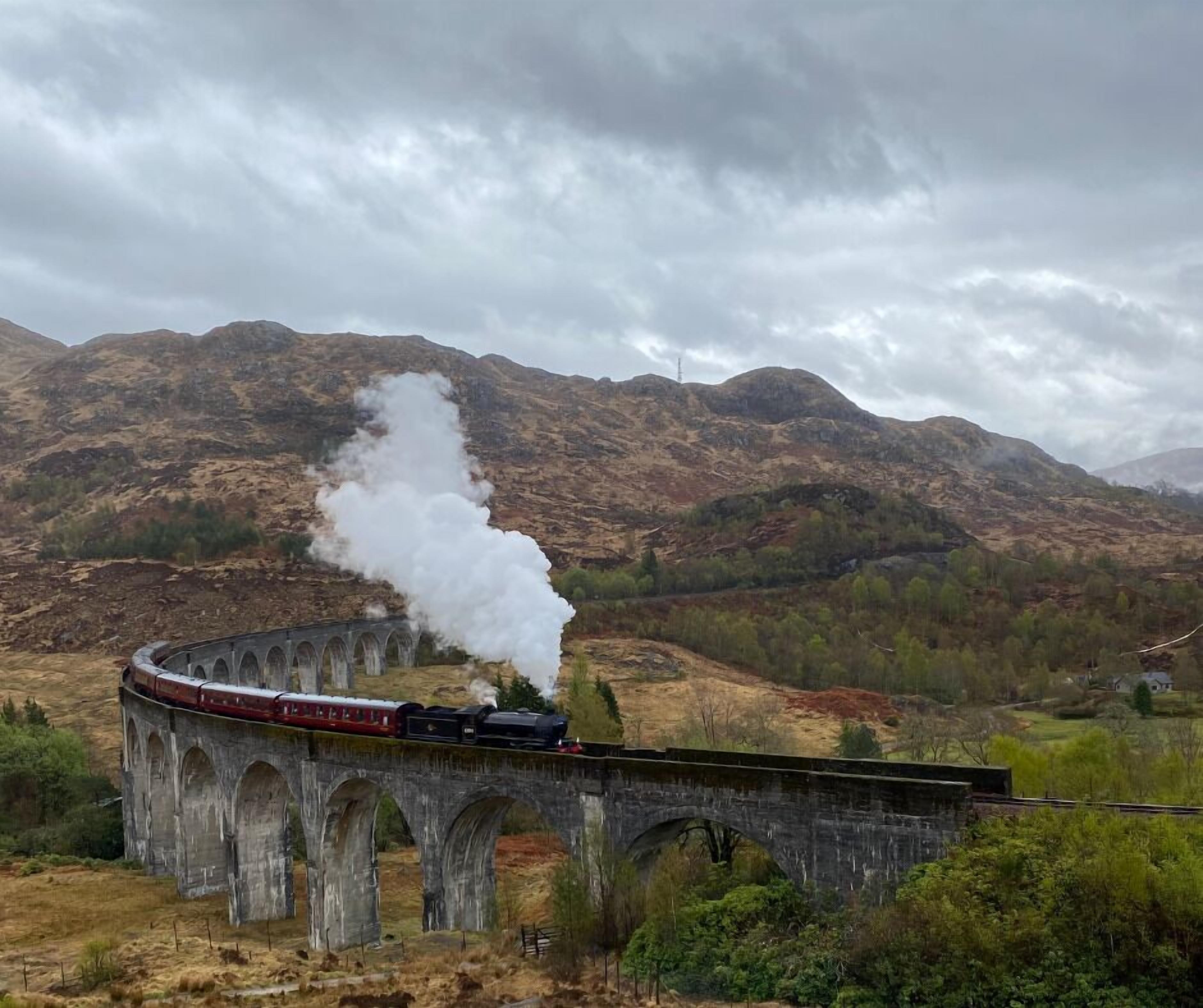 Eine Dampflokomotive überquert ein Steinviadukt in einer Berglandschaft.