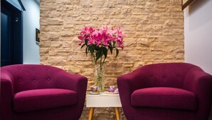 Une pièce avec deux fauteuils violets, une petite table blanche et un vase de fleurs roses, le tout placé devant un mur de pierre.