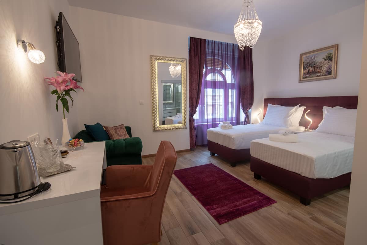 A hotel room with a desk, two beds, a green chair, a red rug, a chandelier, and a painting on the wall.