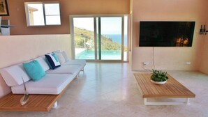Una sala de estar moderna con un sofá blanco, una mesa de centro con una planta, un televisor montado en la pared y un amplio ventanal corredero que da a un balcón con vistas al mar.