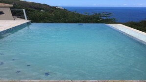 Una piscina infinita rectangular con agua azul cristalina, rodeada por una terraza de piedra, con vistas a un paisaje costero con colinas y el mar.