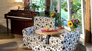 Ein Zimmer mit Klavier, einem armchair im Blumenmuster, einem runden Holztisch und großen Fenstern mit Pflanzen und Blumen.