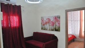 Habitació amb un sofà vermell, un quadre de flors roses, una làmpada al sostre i una porta que dona a una altra habitació.