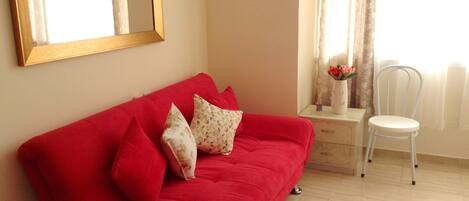 A red sofa with floral cushions, a white chair, a mirror, and a vase with flowers on a small table.