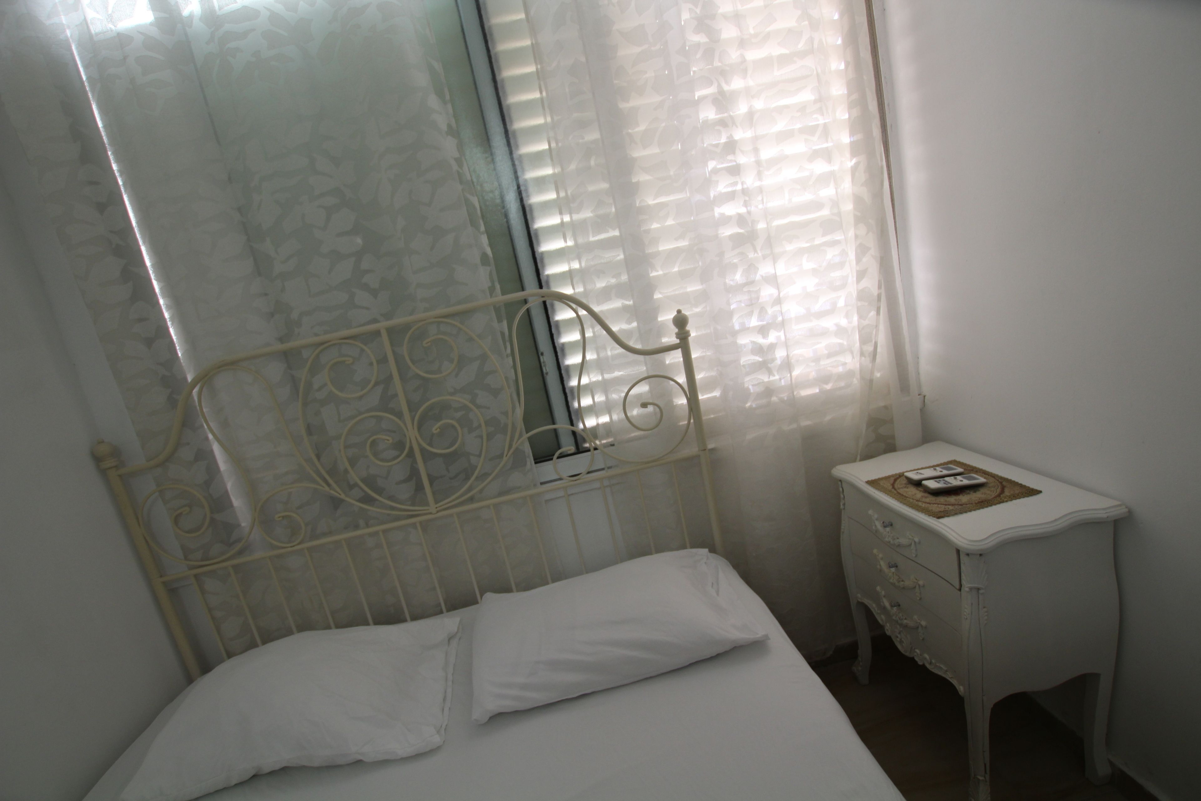 A bedroom with a metal bed frame, white bedding, a bedside table with a remote control, and sheer curtains.