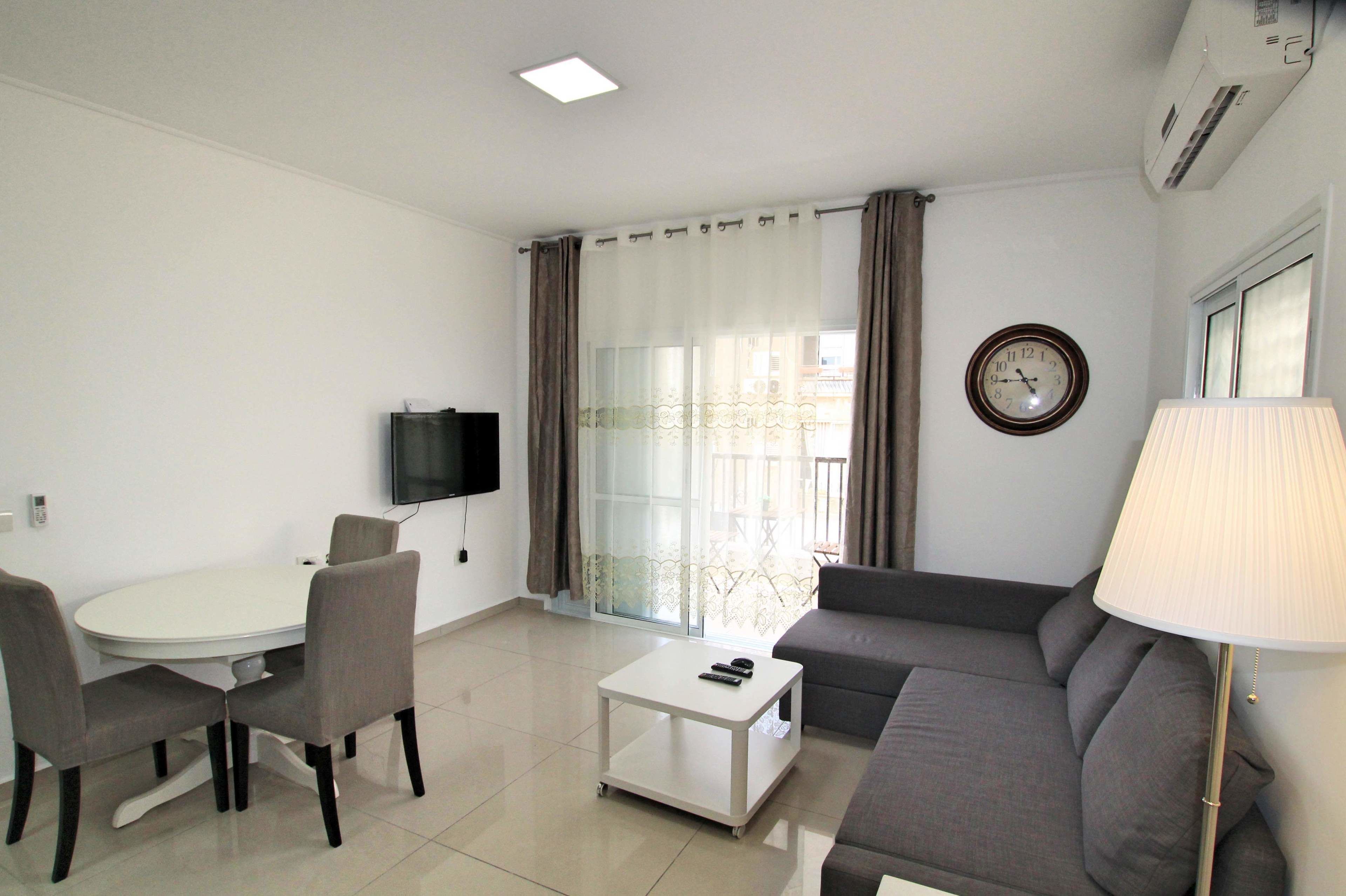 A modern living room with a sofa, a small table, a TV mounted on the wall, and a window with curtains.