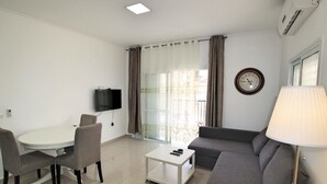 A modern living room with a sofa, a small table, a TV mounted on the wall, and a window with curtains. - Arendalzrail Apartment - Balfour 35 (Bat Yam)