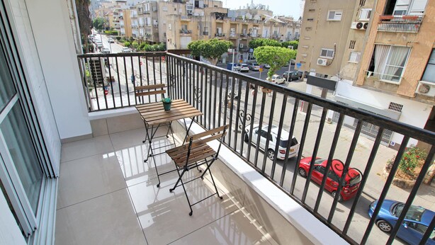 A balcony with a wooden table and two chairs, overlooking a street with parked cars and residential buildings. - Arendalzrail Apartment - Balfour 35 (Bat Yam)