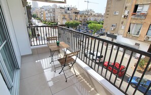 A balcony with a wooden table and two chairs, overlooking a street with parked cars and residential buildings.