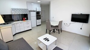 A modern kitchen with white cabinets, a microwave, and a stove. There is a dining area with a table and chairs, and a TV mounted on the wall. - Arendalzrail Apartment - Balfour 35 (Bat Yam)
