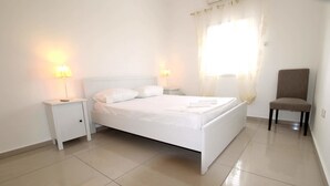 A clean, white bedroom with a bed, two bedside lamps, a chair, and a window with sheer curtains. - Arendalzrail Apartment - Balfour 35 (Bat Yam)