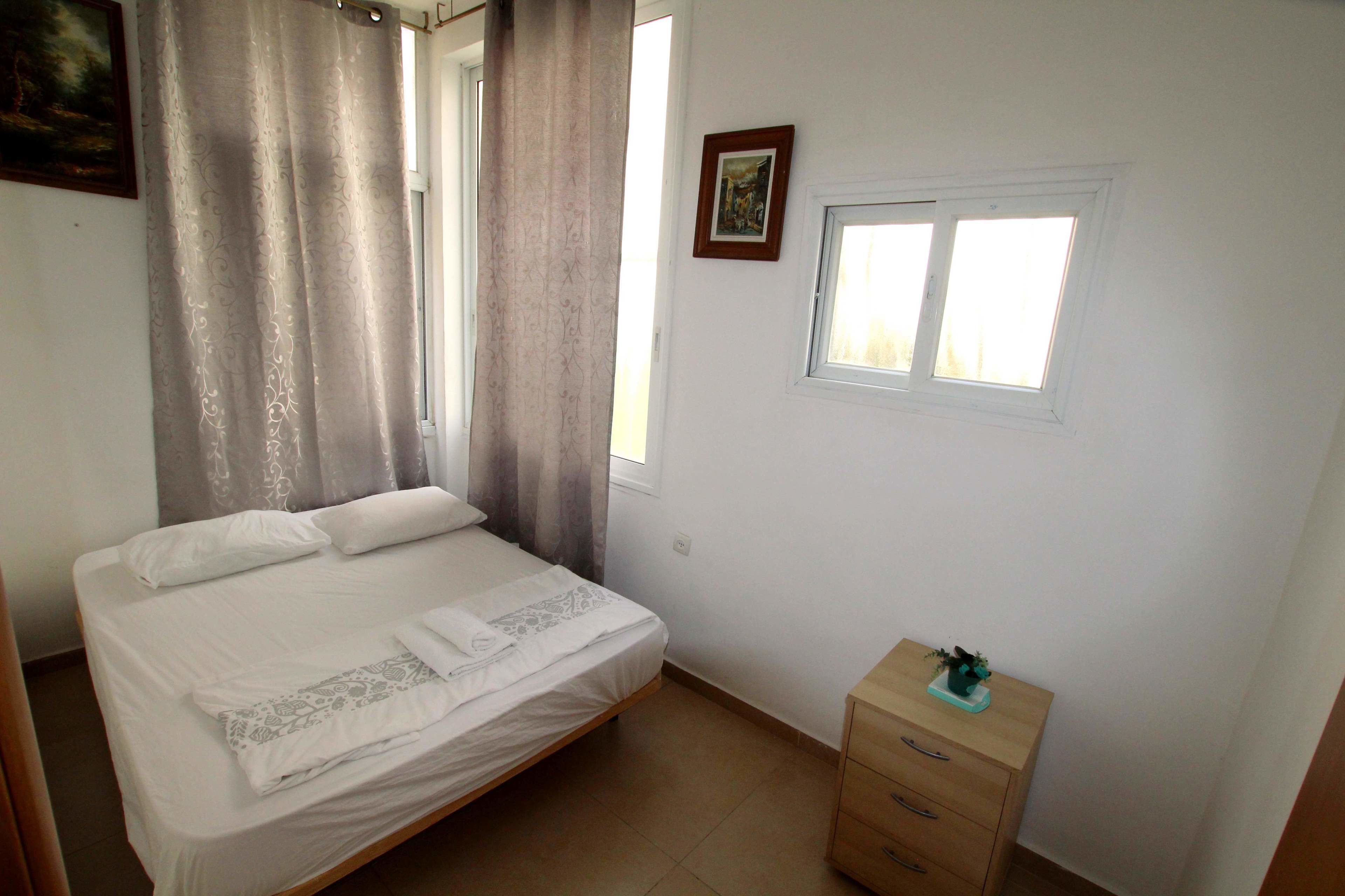 A bedroom with a bed, a window, a small cabinet, and a painting on the wall.