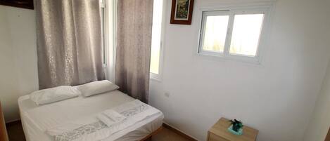 A bedroom with a bed, a window, a small cabinet, and a painting on the wall.