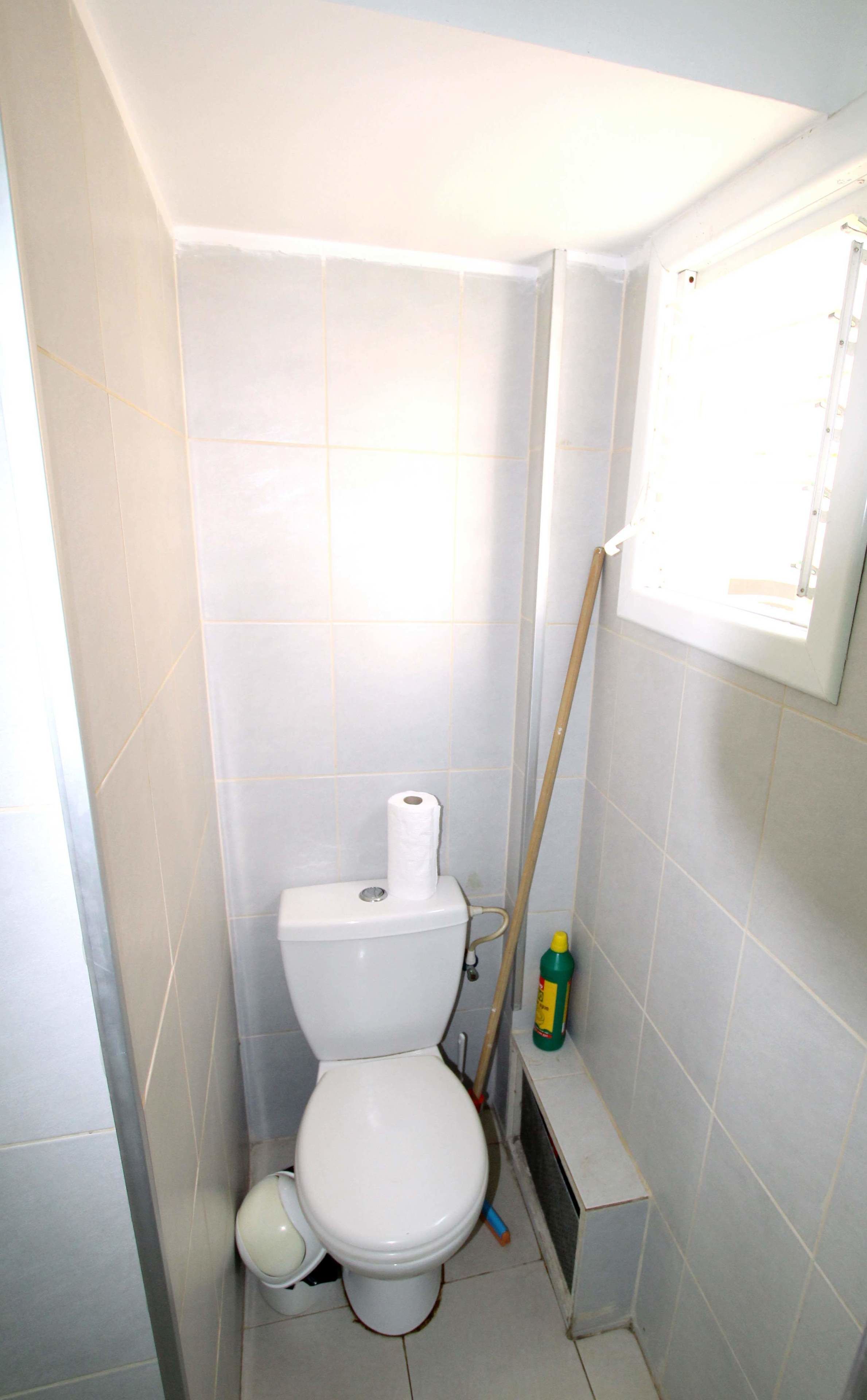 A small, narrow bathroom with a toilet, a roll of toilet paper, a broom, and a cleaning spray bottle.