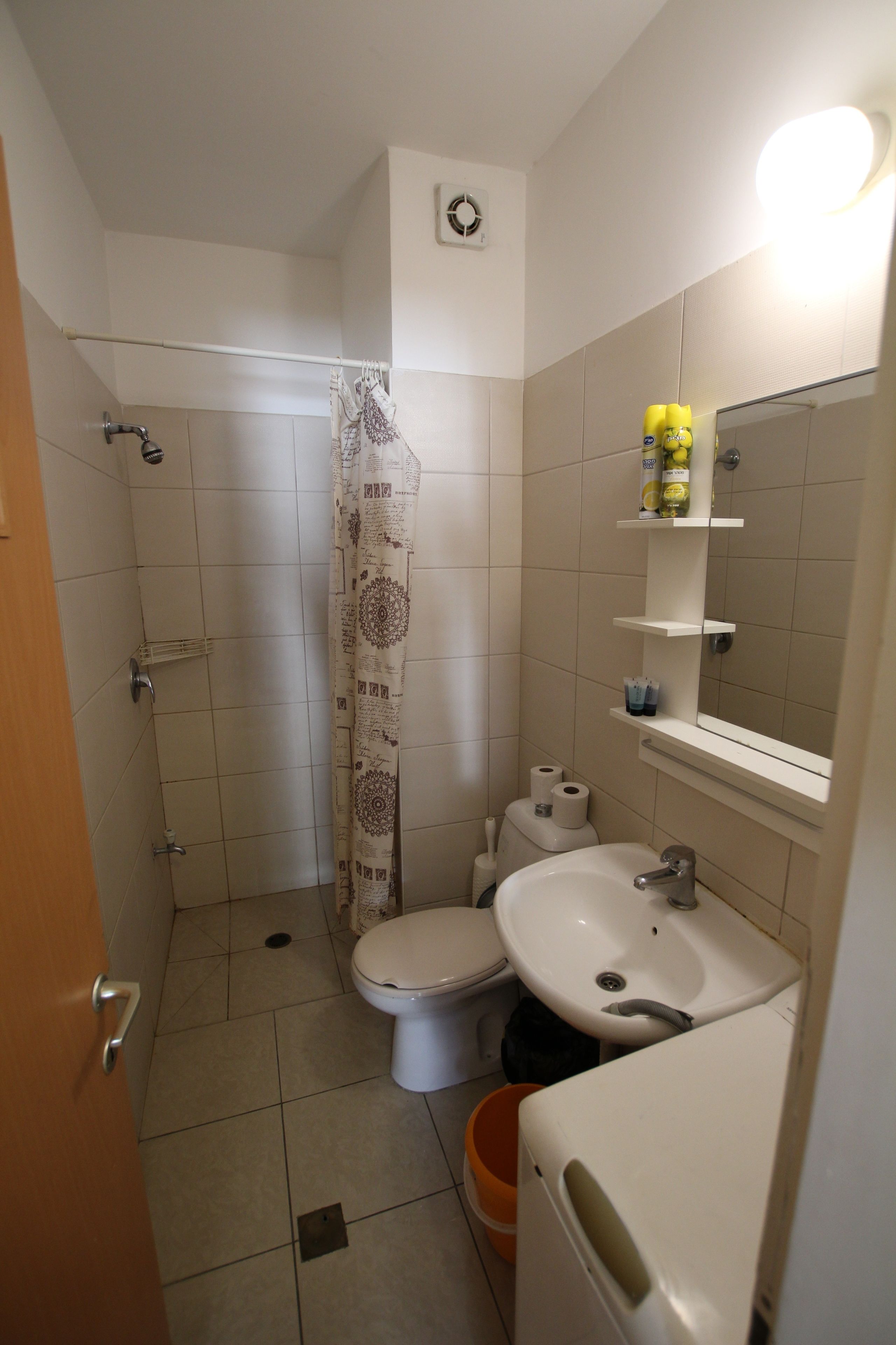 Un baño con inodoro, lavabo y ducha. Hay estantes con artículos de aseo y un espejo sobre el lavabo.