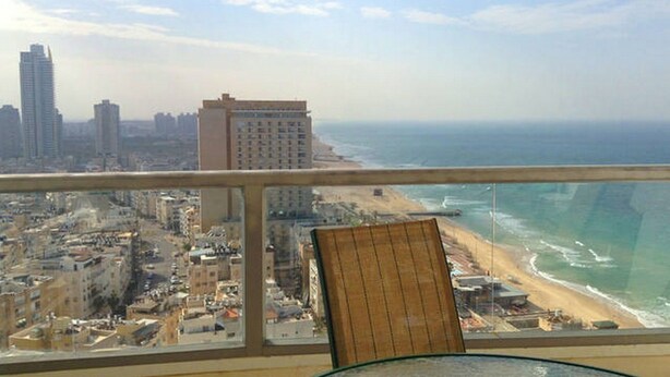 A balcony with a view of a beach and cityscape, including high-rise buildings and a chair. - Arendalzrail Apartments - Yoseftal Street 3 (Bat Yam)
