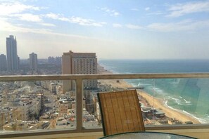 A balcony with a view of a beach and cityscape, including high-rise buildings and a chair. - Arendalzrail Apartments - Yoseftal Street 3 (Bat Yam)