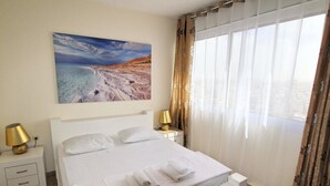 A neatly made bed with white linens and towels, a painting of a beach scene on the wall, and a window with curtains. - Arendalzrail Apartments - Yoseftal Street 3 (Bat Yam)