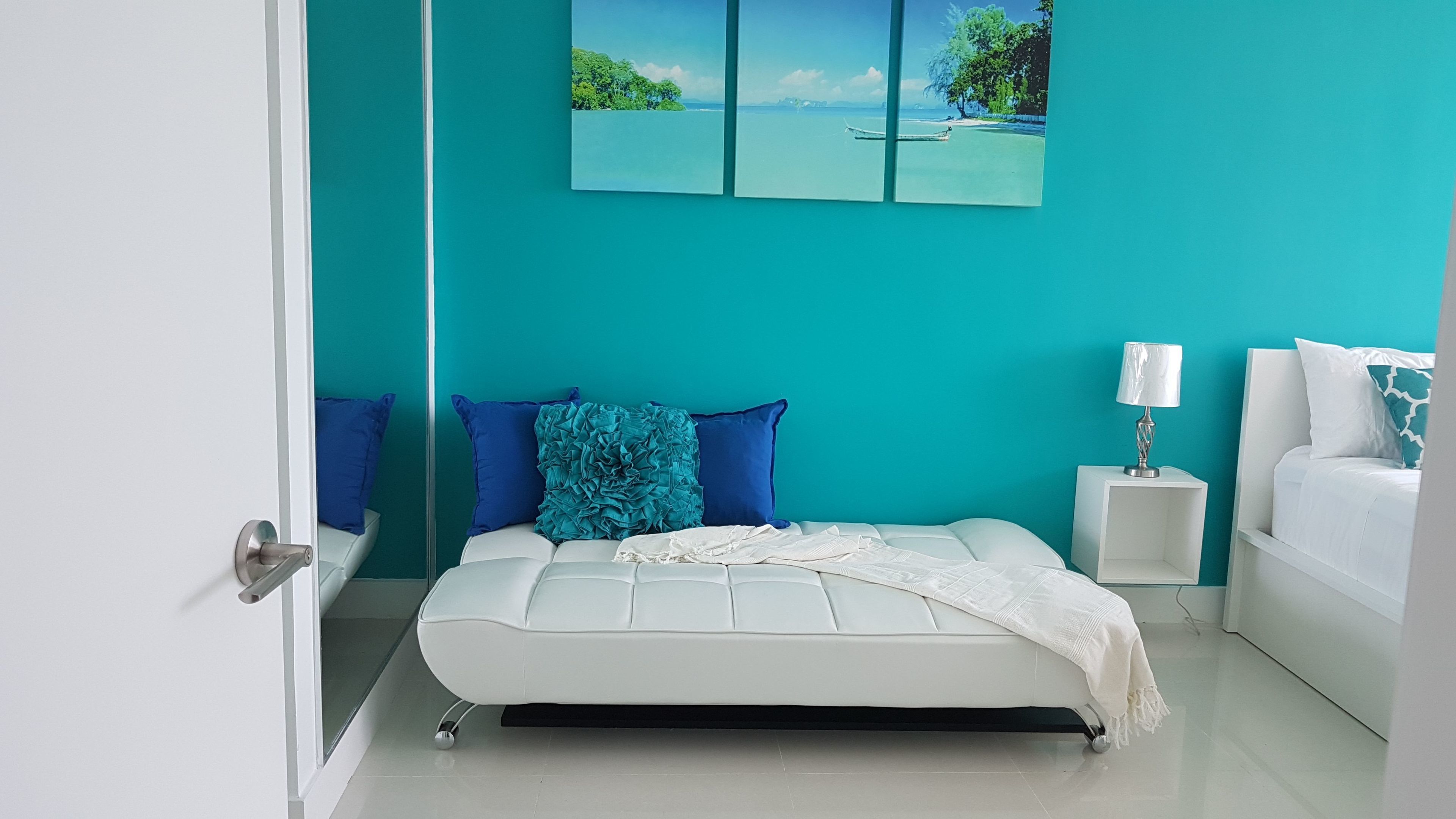 A modern bedroom with a white bed, teal walls, and a large window with a view of a beach and ocean.