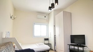 Una habitación pequeña y luminosa con cama, sofá, televisor sobre un mueble bajo, ventana y armario.