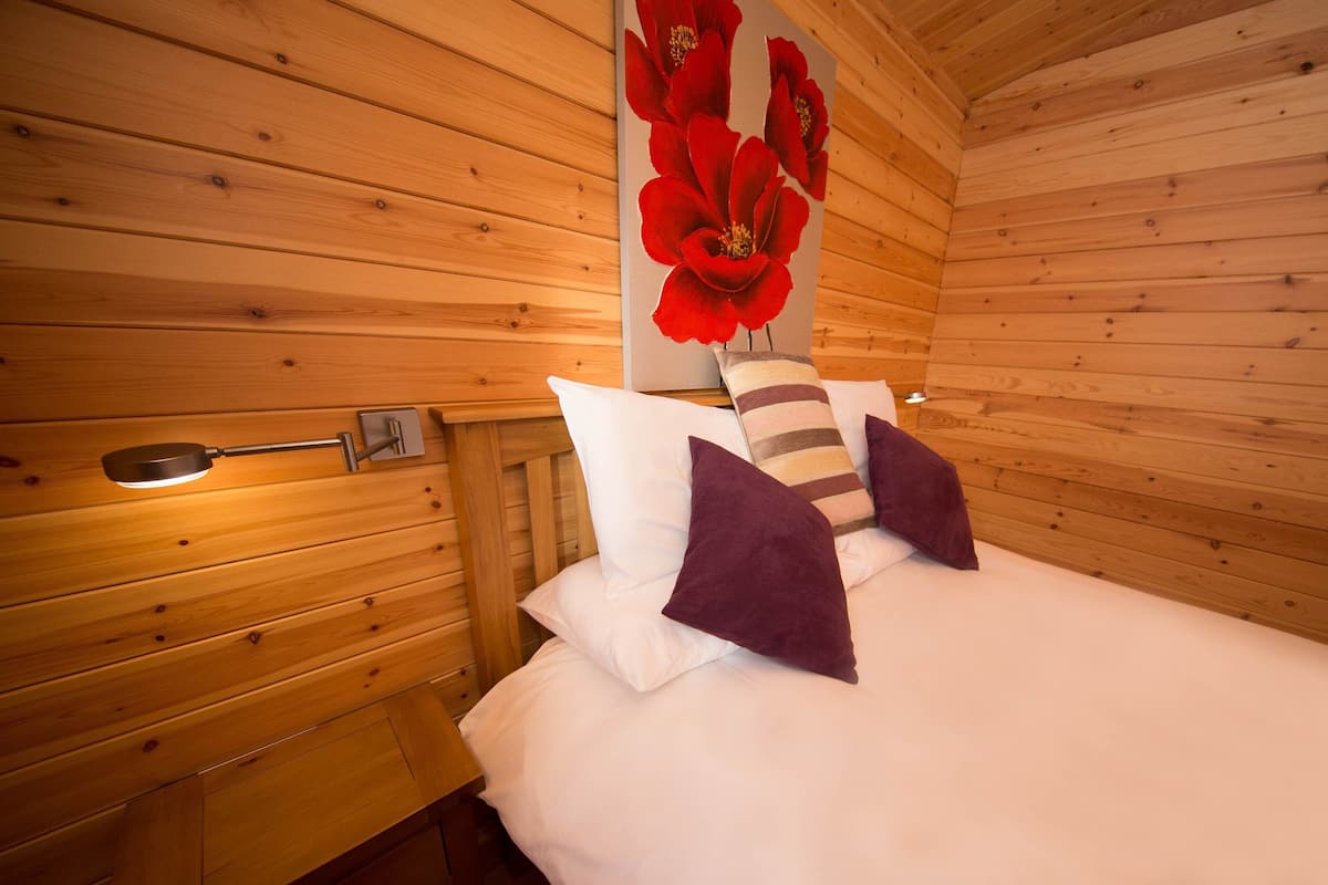 Una camera di una baita in legno con un letto, cuscini, un comodino e una lampada fissata alla parete.
