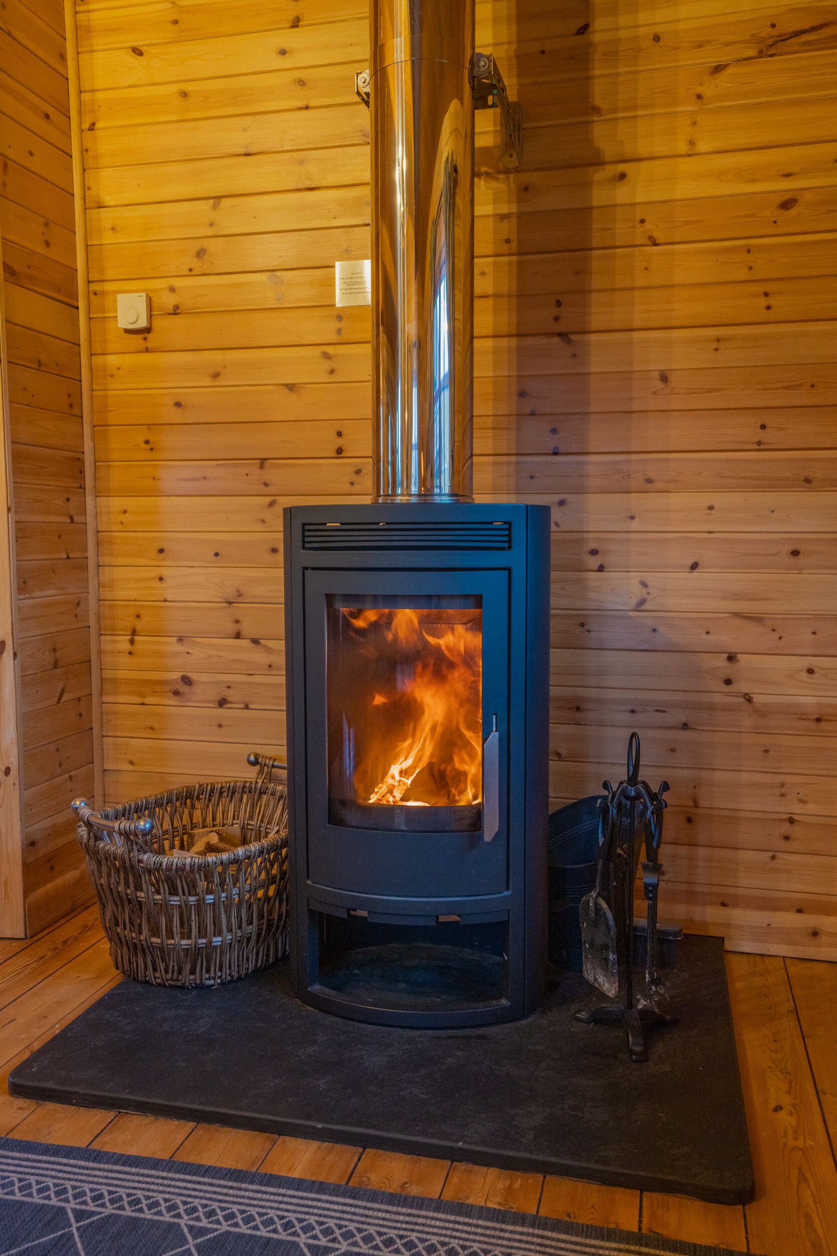 Una estufa de leña con fuego, una cesta tejida y un gancho de metal sobre un felpudo en una cabaña de madera.