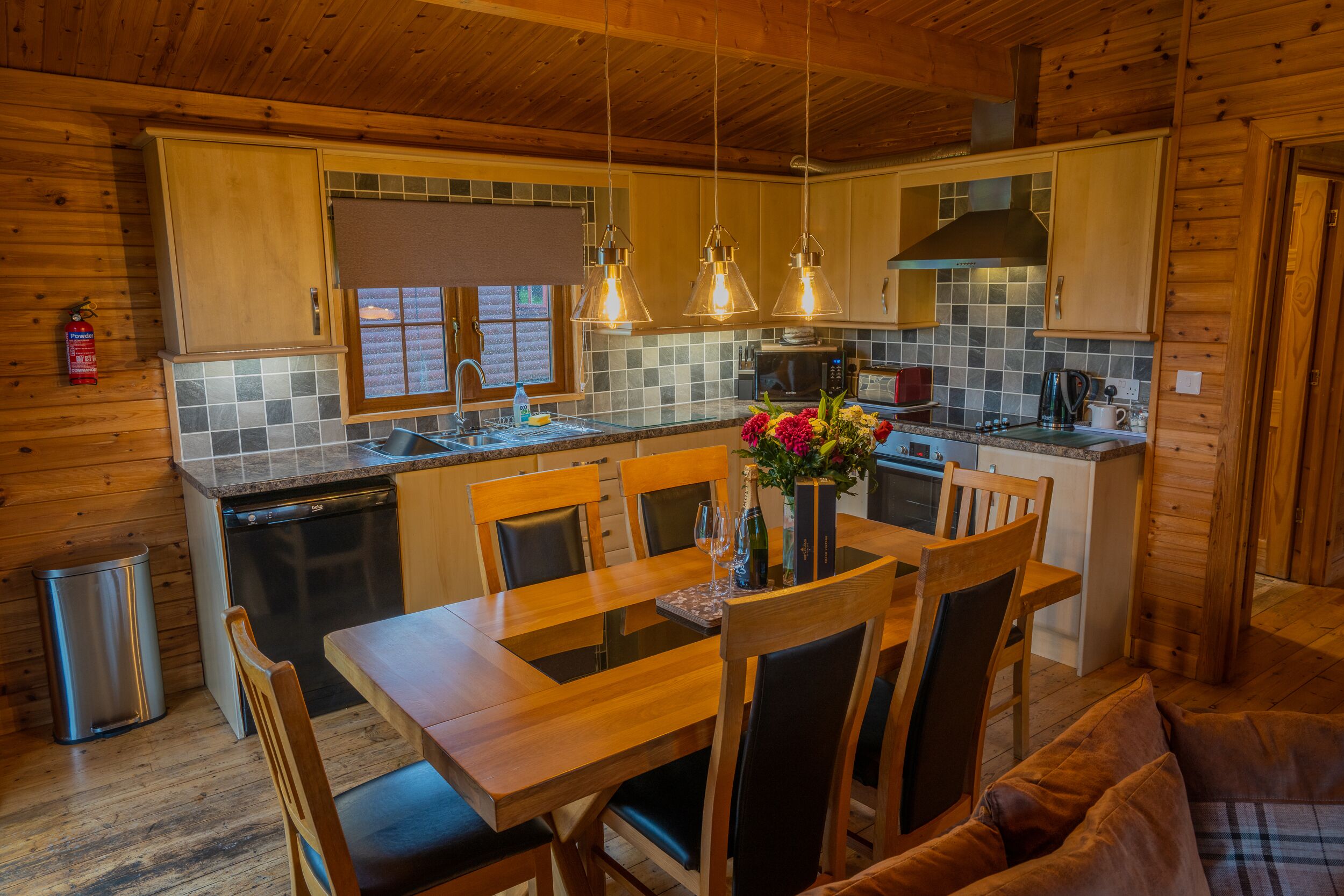 A wooden cabin interior with a dining table, chairs, a kitchen area with a microwave, and a fire extinguisher.