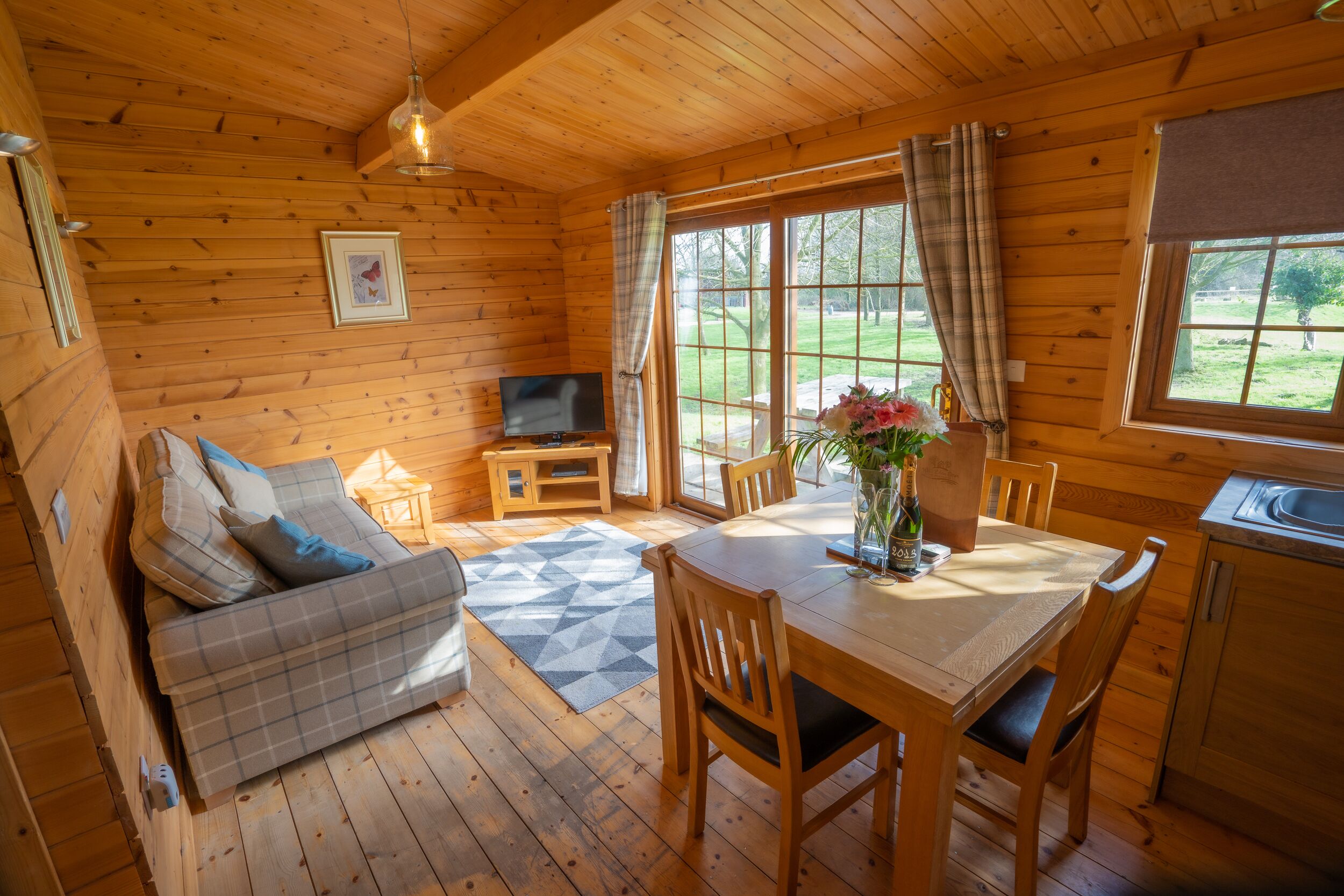 A cozy cabin interior with a wooden floor, a sofa, a dining table with chairs, a television, and a window with curtains.