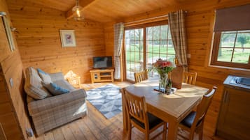 A cozy cabin interior with a wooden floor, a sofa, a dining table with chairs, a television, and a window with curtains.