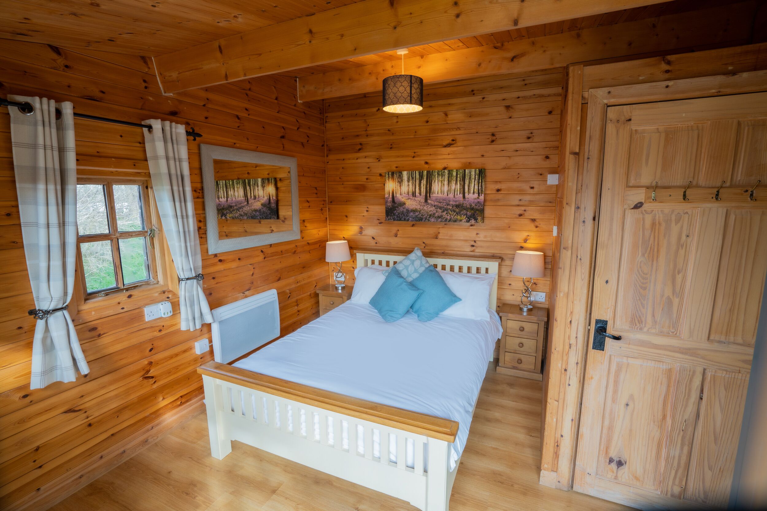 Ein Schlafzimmer einer Holzhütte mit einem Bett, zwei Nachttischen, einem Schminktisch, einem Fenster mit Vorhängen und einer Tür.