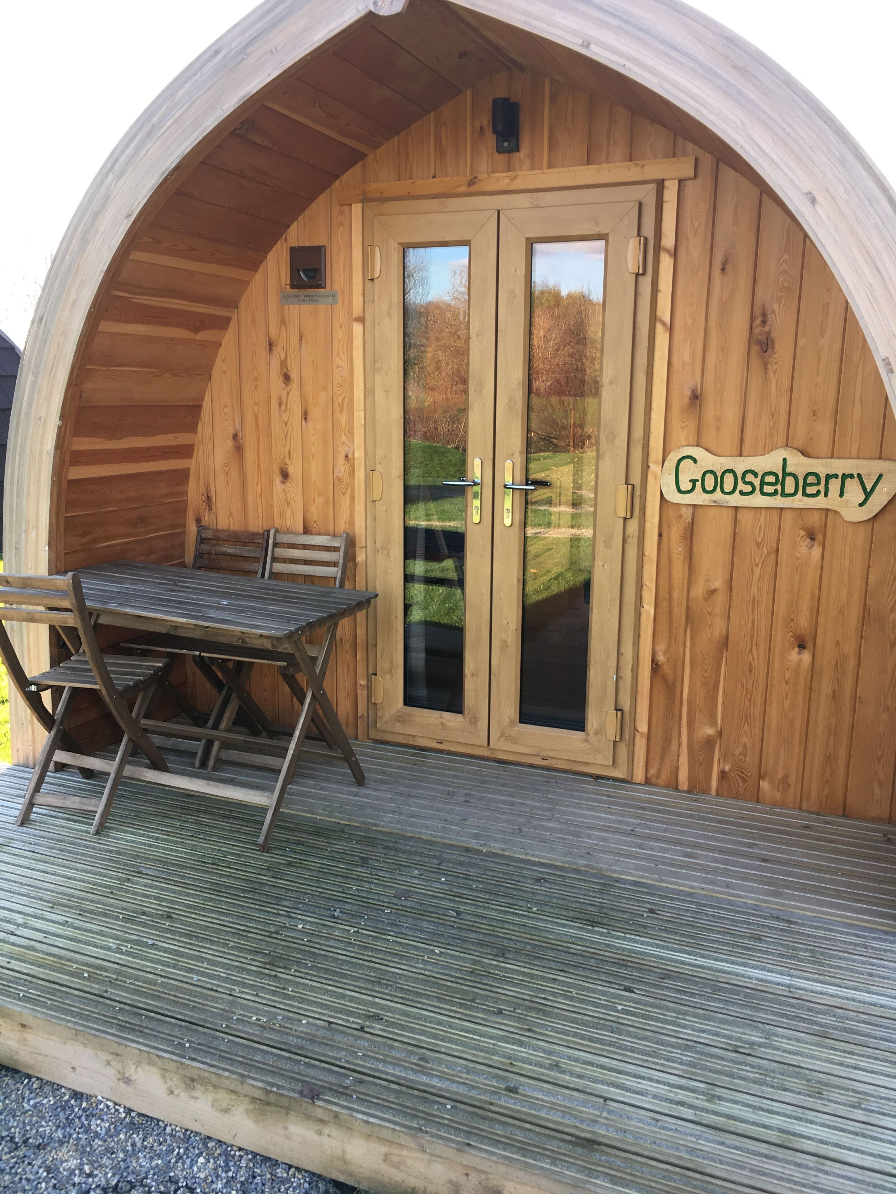 En træhytte med et bord og stole udenfor, et skilt med teksten "Gooseberry" og udsigt over et grønt landskab gennem døren.