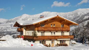 A multi-story wooden chalet with balconies, set against a snowy mountain backdrop.