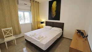 A neatly made bed with white linens, a wooden nightstand with a lamp, a chair, and a feather artwork on the wall. - Arendalzrail Apartments Khana Szenes St. (Bat Yam)