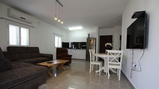 A modern living room with a brown sofa, a wooden coffee table, and a white dining table with chairs. There is a kitchen area with white cabinets and a refrigerator. A flat-screen TV is mounted on the wall. - Arendalzrail Apartments Khana Szenes St. (Bat Yam)