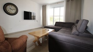 A living room with a brown leather sofa, a wooden coffee table, a brown armchair, a flat-screen TV mounted on the wall, and a window with curtains. - Arendalzrail Apartment - Hertsel St. 4 (Bat Yam)