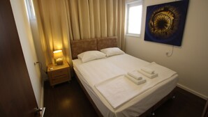 A hotel room with a neatly made bed, a bedside lamp, a nightstand, and a wall-mounted air conditioner. - Arendalzrail Apartment - Hertsel St. 4 (Bat Yam)