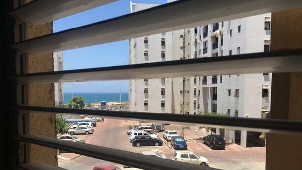 A view from a window overlooking a parking lot with several cars, a red car in the foreground, and a building with multiple floors and balconies. - Arendalzrail Apartment - Hertsel St. 4 (Bat Yam)