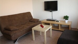 A living room with a brown sofa, a small wooden table, a TV on a stand, a plant, and a framed picture on the wall.
