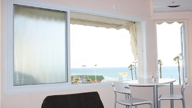 A room with a large window offering a view of the beach and ocean. There is a round table with two chairs and a dark-colored sofa.
