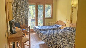 Una habitación con dos camas, un escritorio con ordenador, una silla y una ventana con vistas a árboles.
