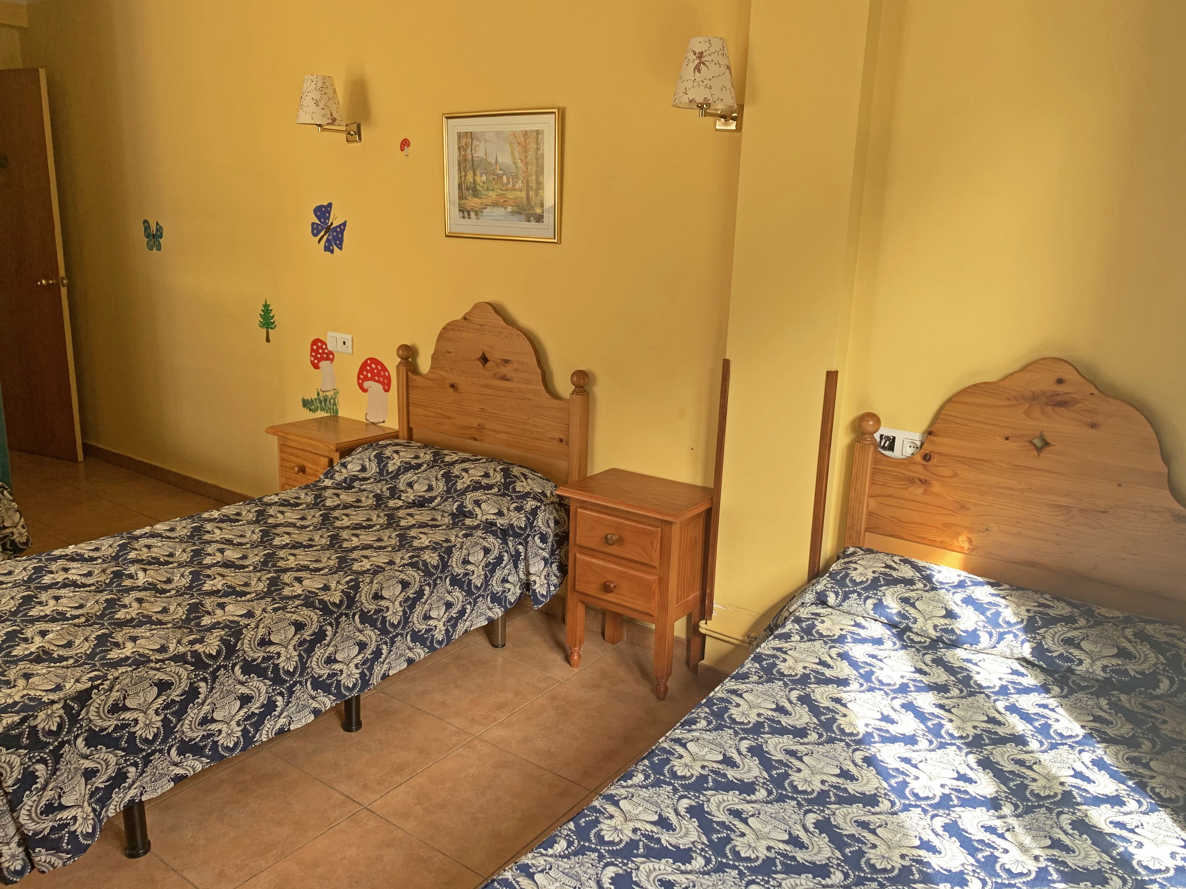 A bedroom with two single beds, wooden headboards, a nightstand, a dresser, and a wall-mounted lamp.