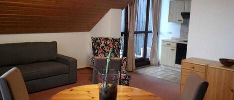 A cozy living area with a wooden ceiling, a grey sofa, a floral armchair, a round wooden table with a glass, and a kitchen in the background.
