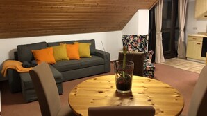 Ett mysigt vardagsrum med ett träpanelat tak, en grå soffa med orange och gröna kuddar, ett runt träbord med stolar och en liten krukväxt.