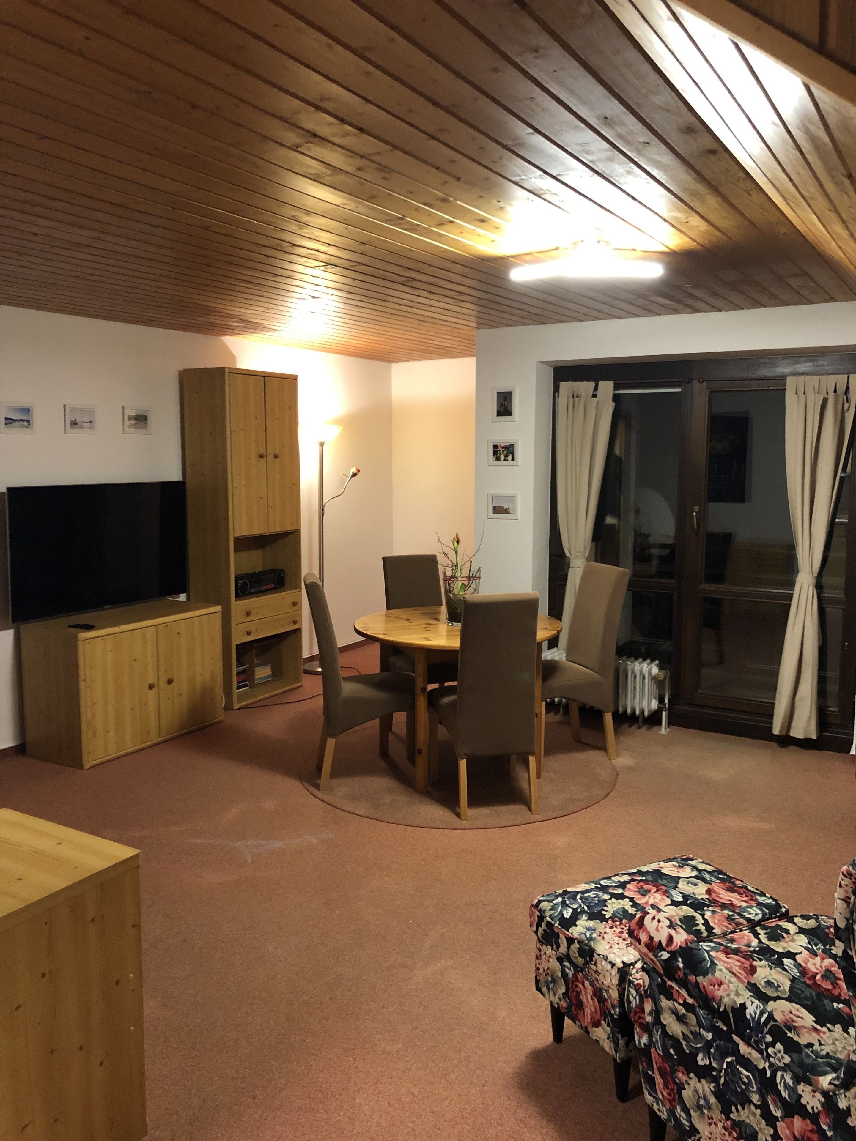 A cozy living room with a wooden ceiling, a TV, a wooden cabinet, a dining table with chairs, and a floral ottoman.