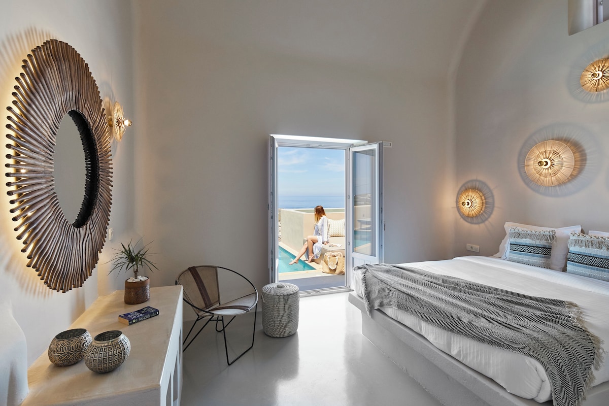 Un dormitorio con una cama grande, vistas al mar a través de una puerta abierta, un espejo redondo y una pequeña mesa con adornos.
