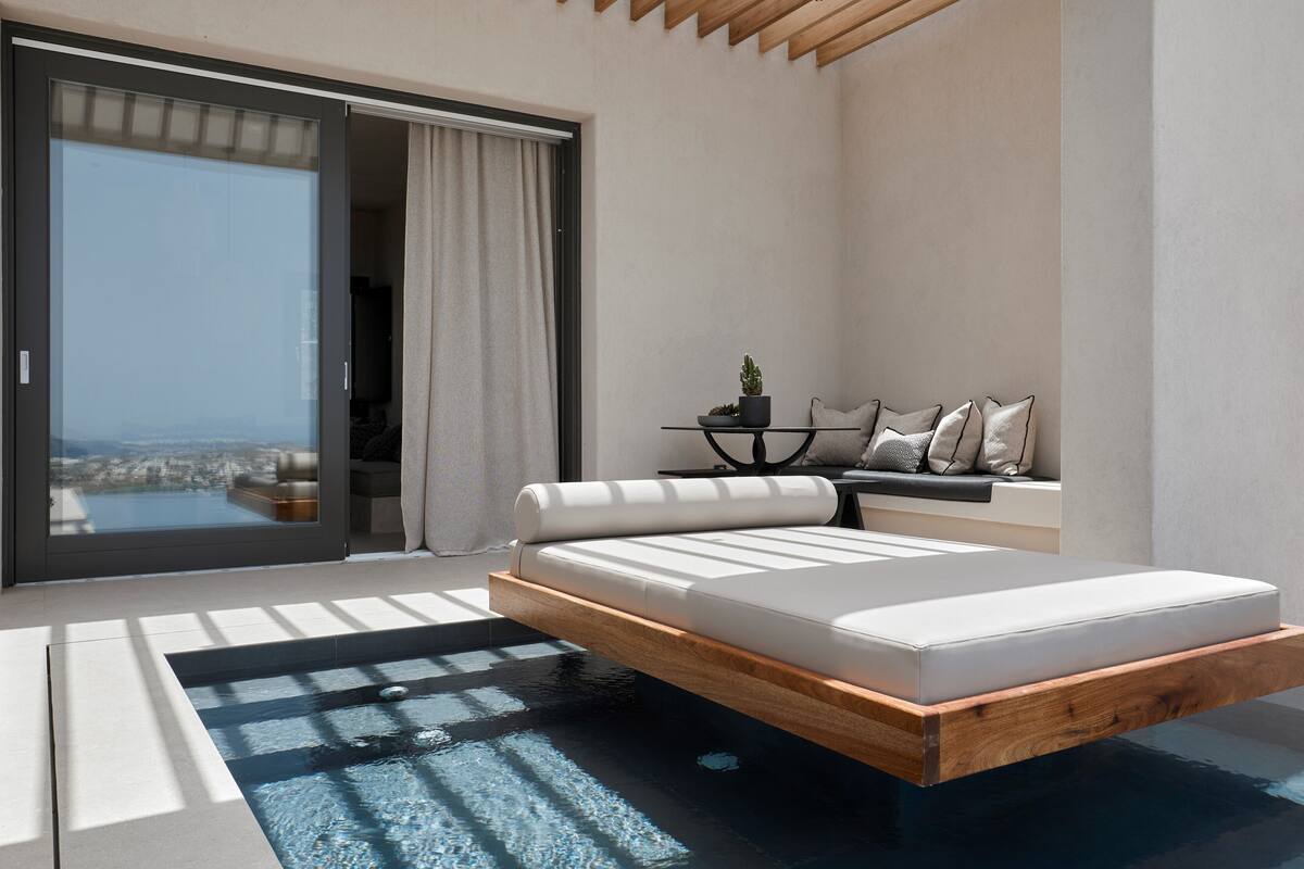 Une chambre moderne avec un grand lit, une petite table ornée d’une plante et une vue sur la ville, visible à travers une porte coulissante.