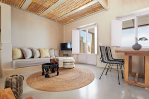A modern living room with a sofa, coffee table, and chairs, a television, and a large window with blinds. - North Santorini A Luxury Spa Hotel (Santorini)