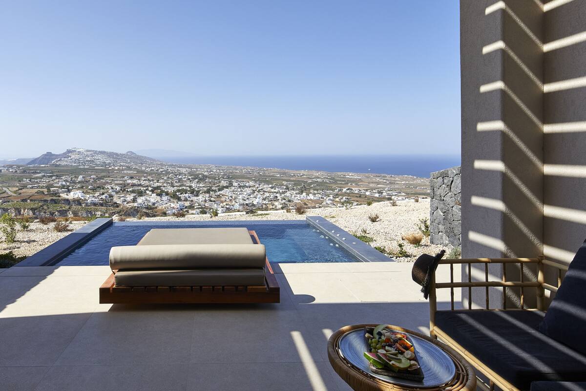 Un espace aménagé au bord de la piscine, avec vue sur une ville côtière, comprenant un fauteuil de relaxation et une table à manger avec une assiette de nourriture.