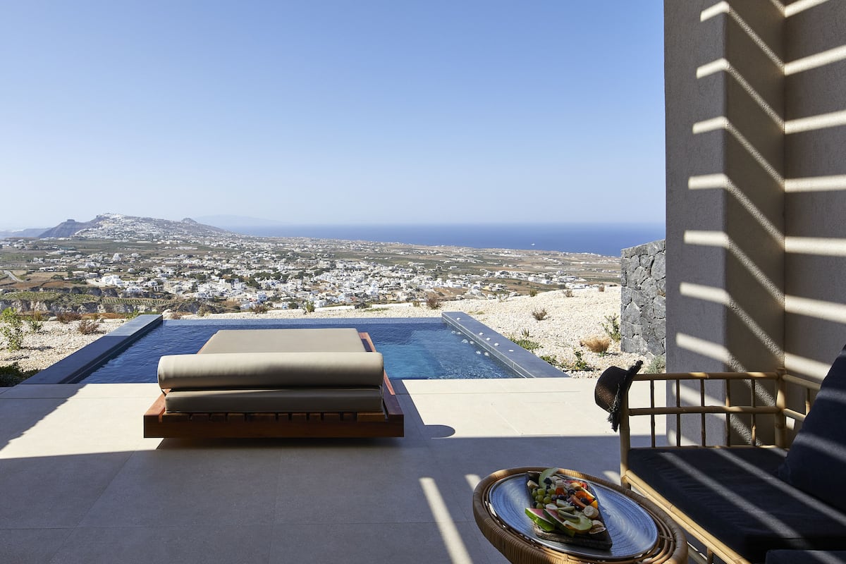 Un espace aménagé au bord de la piscine, avec vue sur une ville côtière, comprenant un fauteuil de relaxation et une table à manger avec une assiette de nourriture.